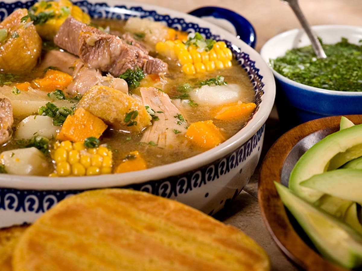 Sancocho llanero cruzado servido en una olla de barro con carne, gallina, verduras y mazorca, acompañado de arepas y aguacate